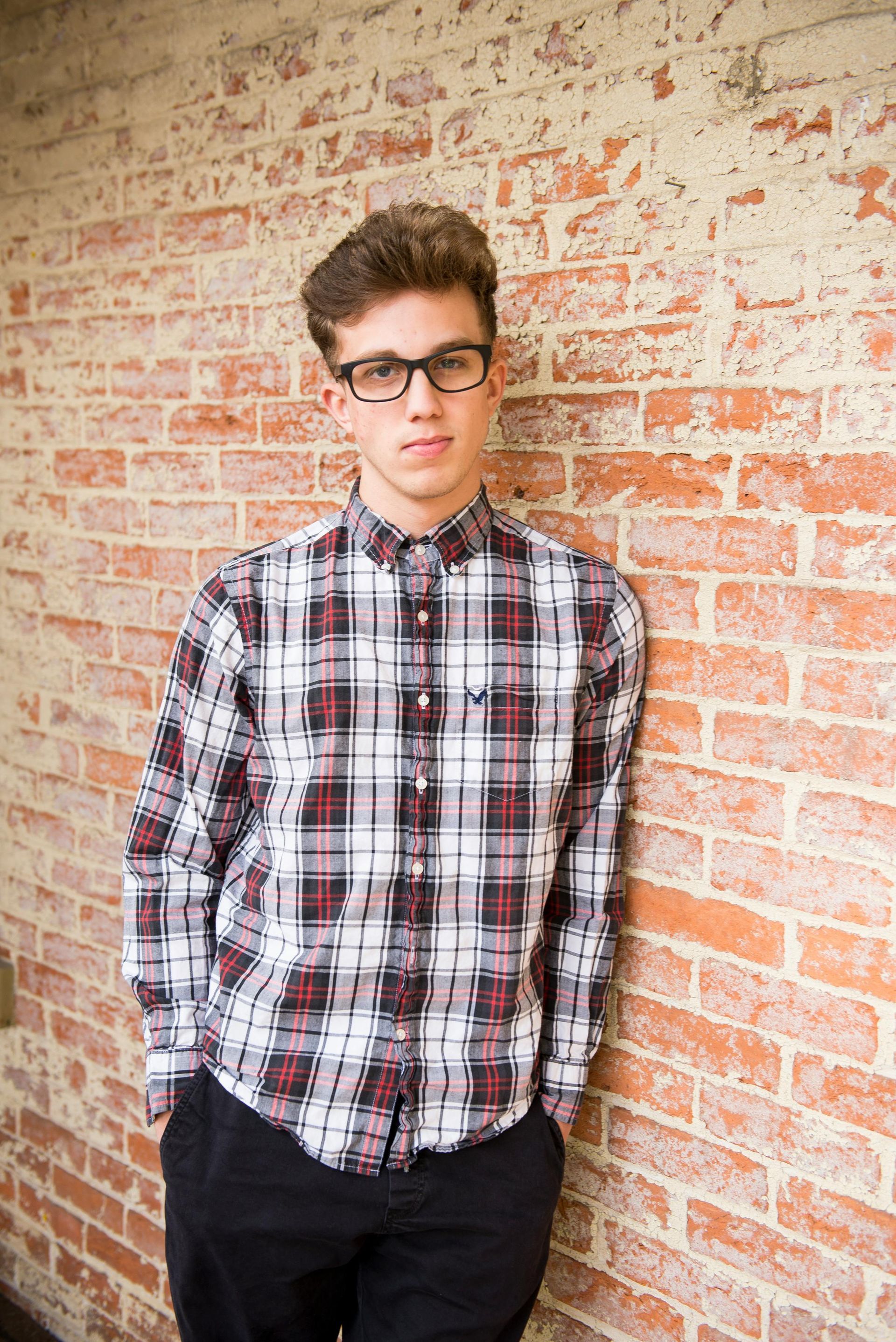 A young man wearing glasses and a plaid shirt leans against a brick wall