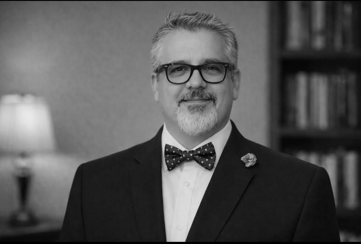 Man in suit and bow tie, wearing glasses, smiling slightly, in front of a bookshelf.
