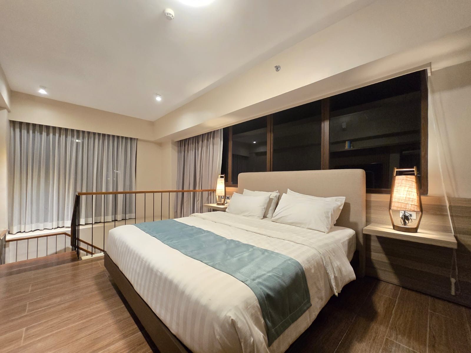 Modern bedroom with a neatly made bed, wood flooring, large window, and warm bedside lamp lighting