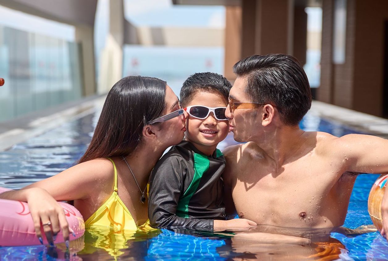 Family in a swimming pool, kissing a child. The pool has blue tiles.