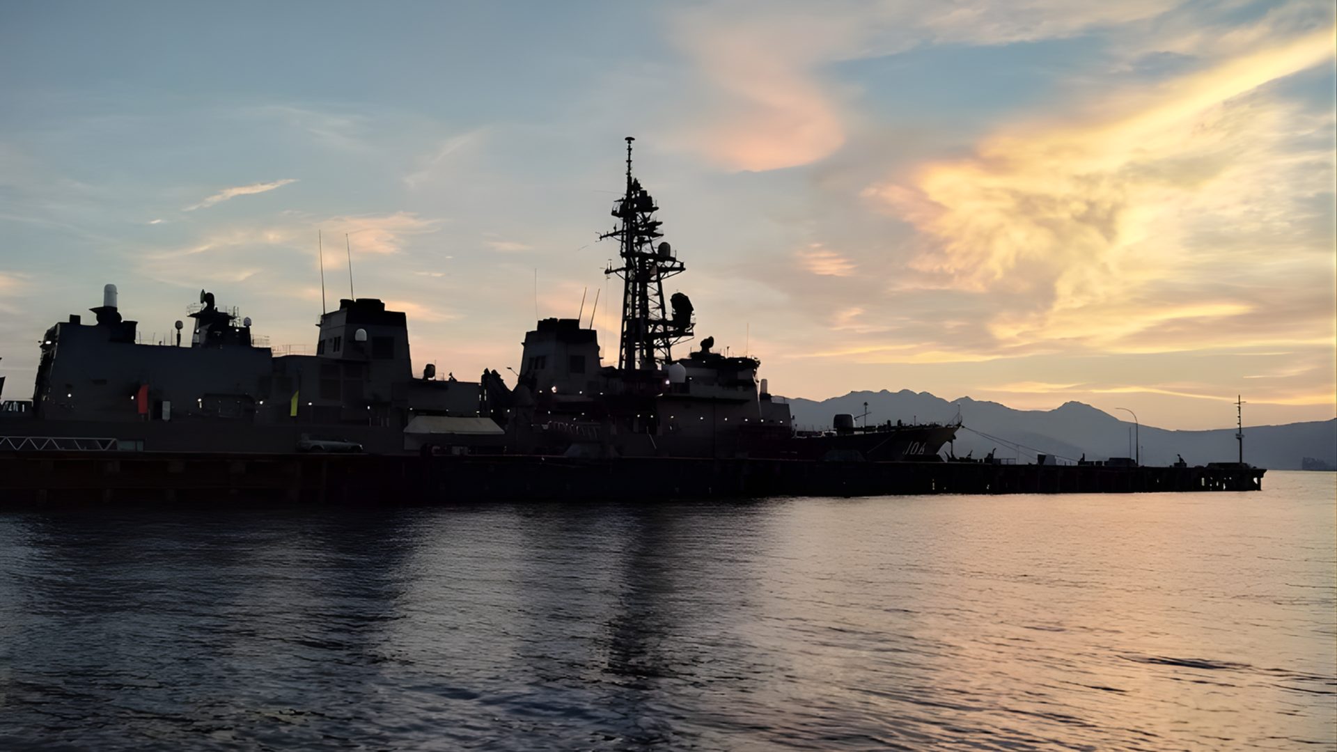 Silhouetted naval ship at sunset on calm water, with mountains in the distance