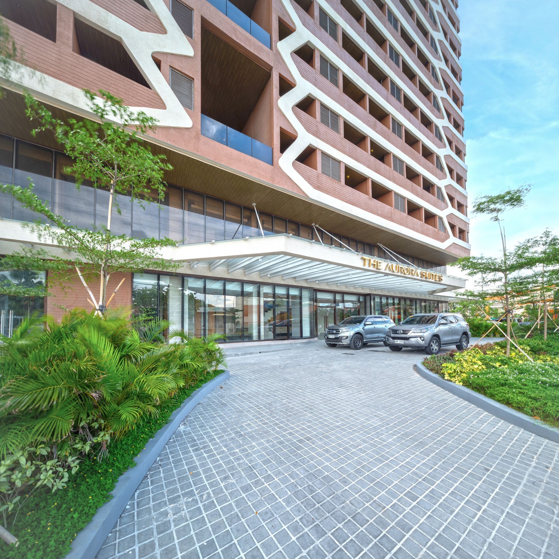 Modern apartment tower entrance with parked cars, curved driveway, and landscaped greenery