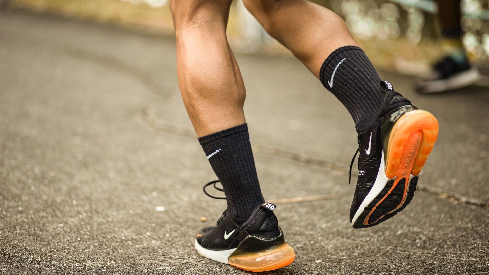 Runner's legs and feet in black Nike sneakers with orange soles, black socks, running on a gray path.