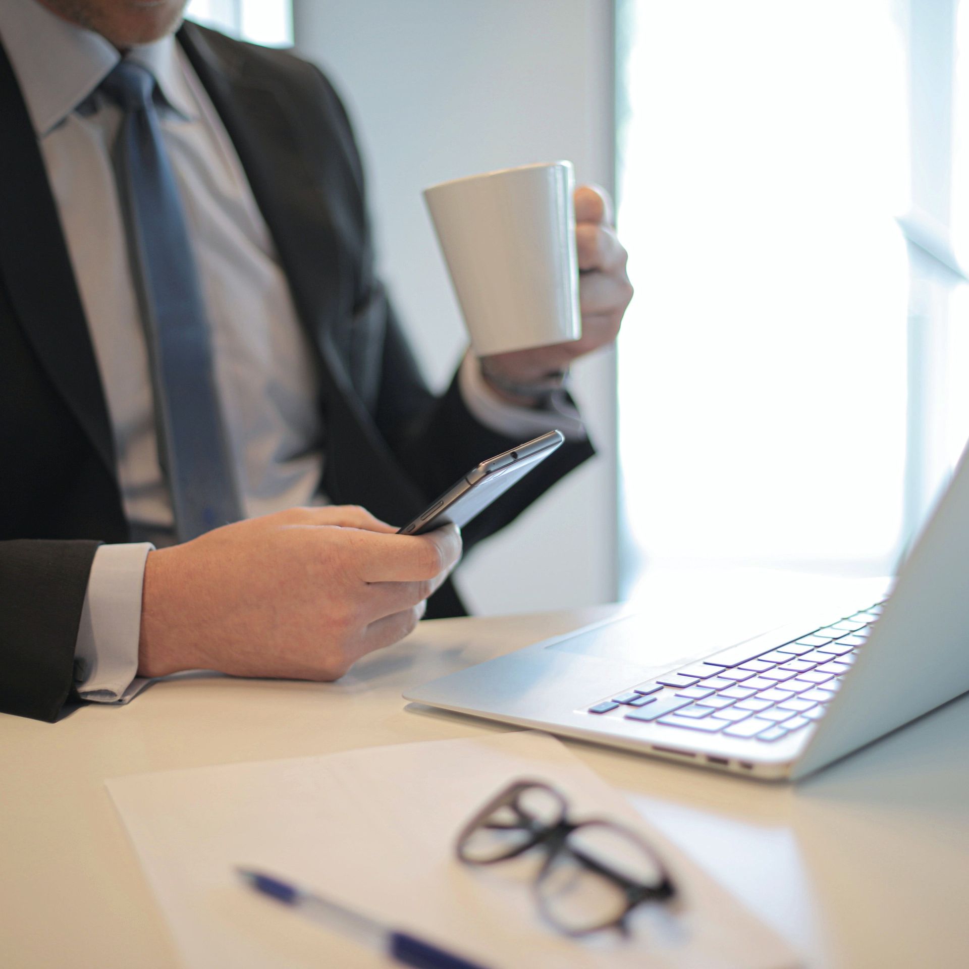 A person in a suit sits at a desk, holding a coffee mug and a smartphone next to a laptop, pen, and eyeglasses.