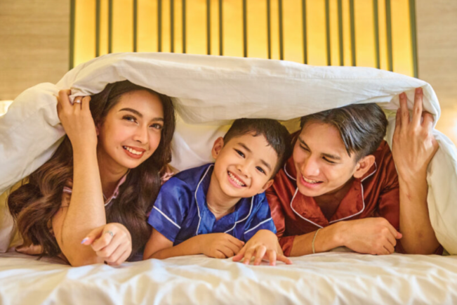 Family, smiling, under a white blanket on a bed.