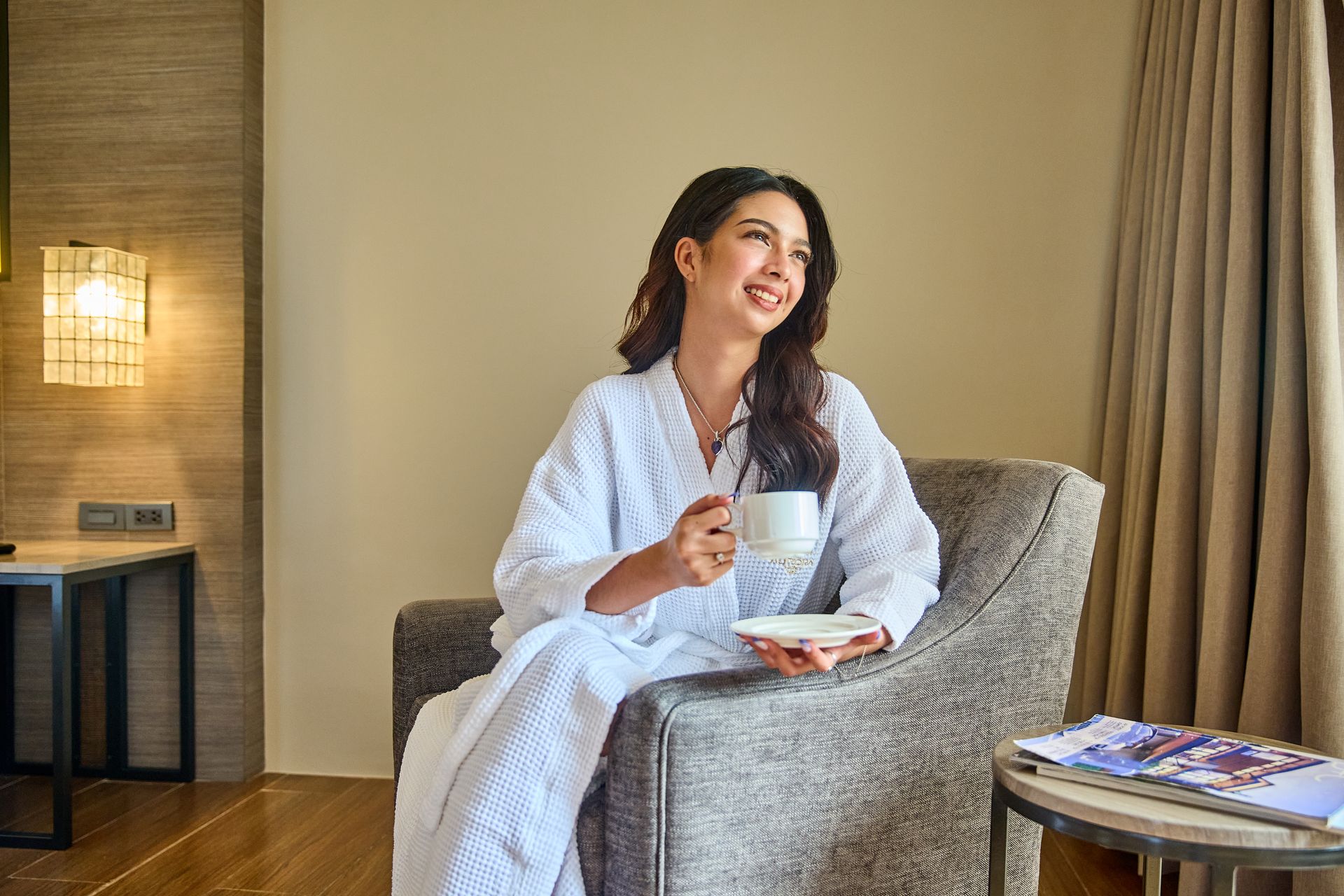 Woman in white robe sitting in chair, holding a cup, smiling near a small table in a room.