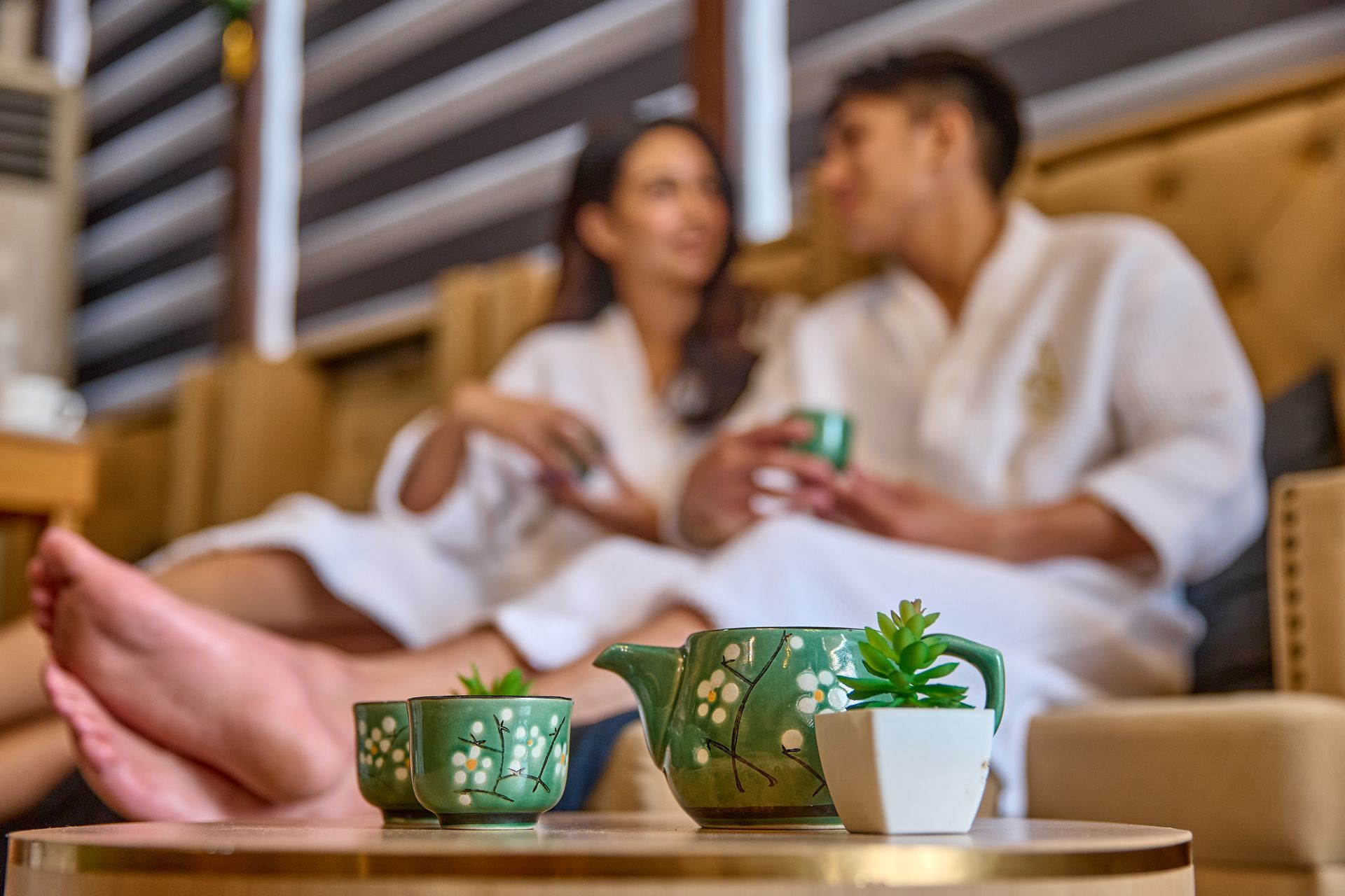 Couple in bathrobes relax, sipping tea in a spa setting. Green teapot and cups on a table.