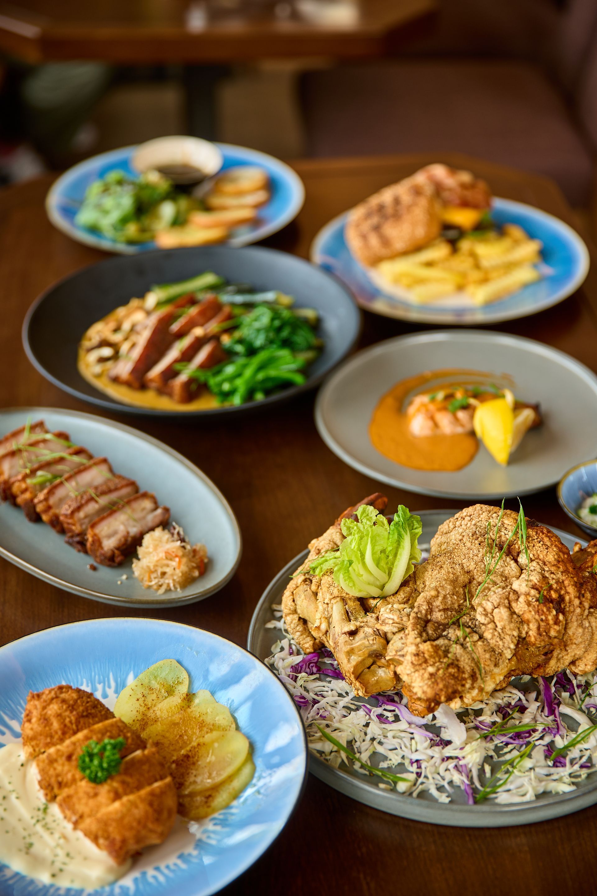 Multiple plates of diverse food on a wooden table. Dishes include fried chicken, fish, and pork with various sides.