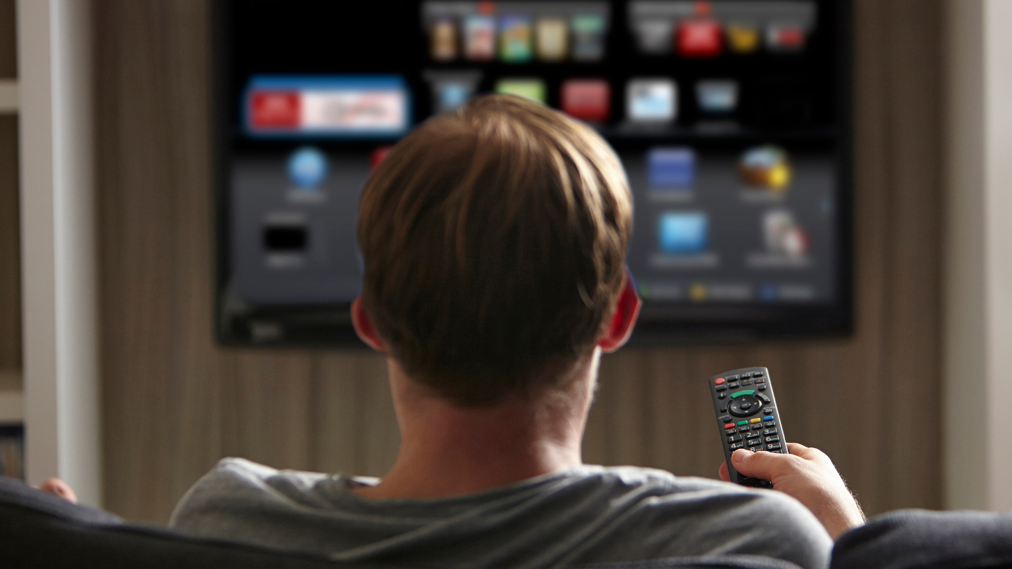 A man is sitting on a couch holding a remote control in front of a television.