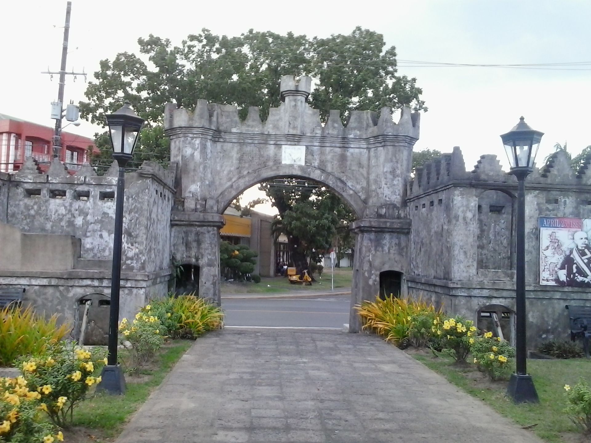 Stone castle entrance with arched gate, lanterns, and flower beds along a paved path