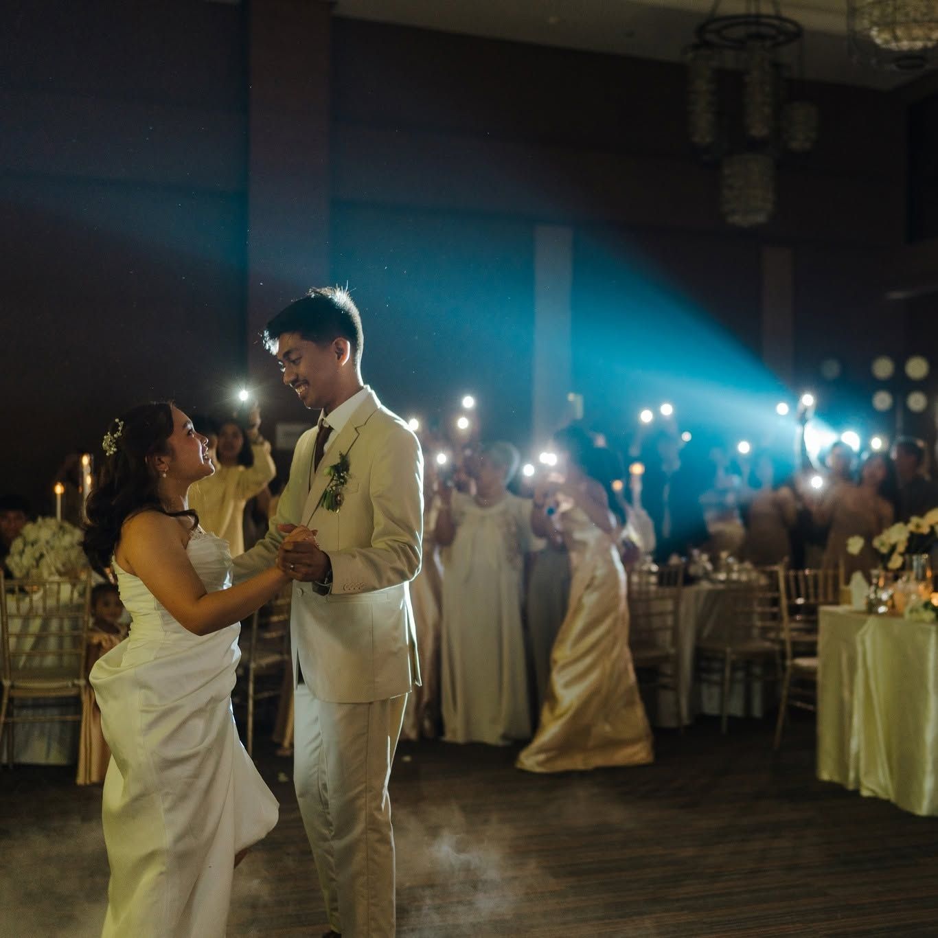 Bride and groom dancing in a dimly lit reception hall as guests hold up phone lights