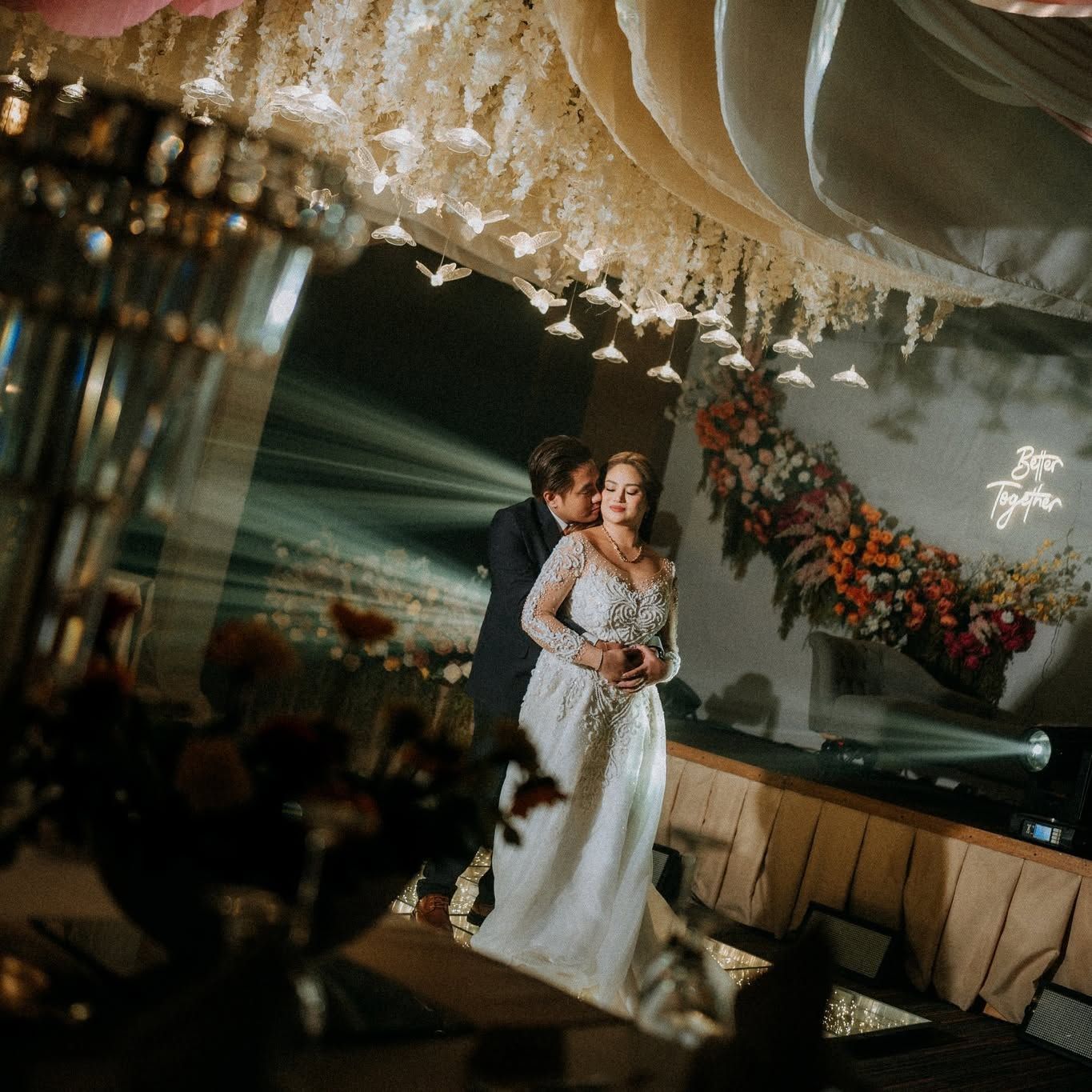 Bride and groom share a dance in a dimly lit reception hall with floral decor and draped ceiling