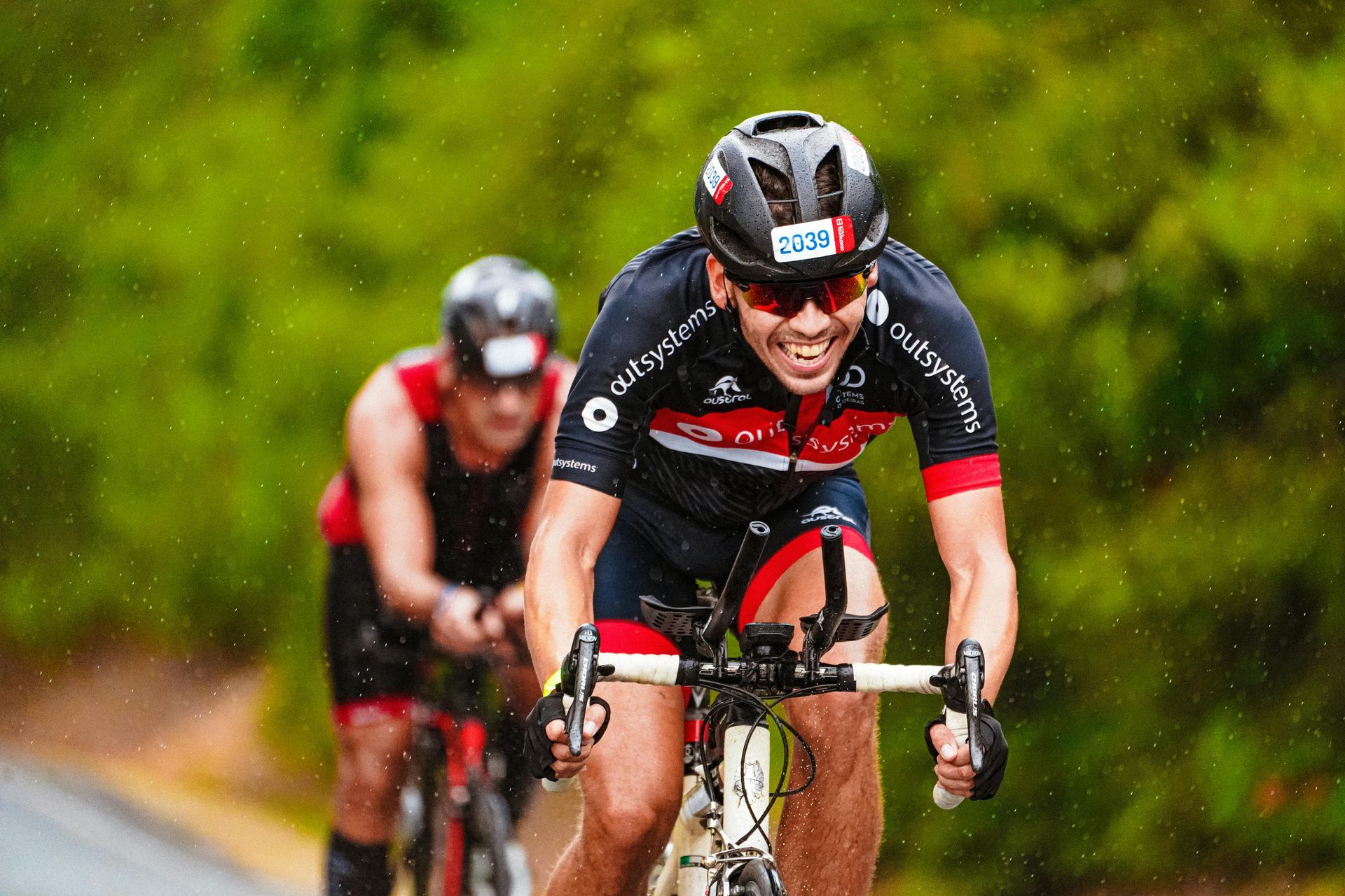 Cyclists in rain: One in focus, smiling, wearing helmet and sunglasses. Another cyclist in background on a road.