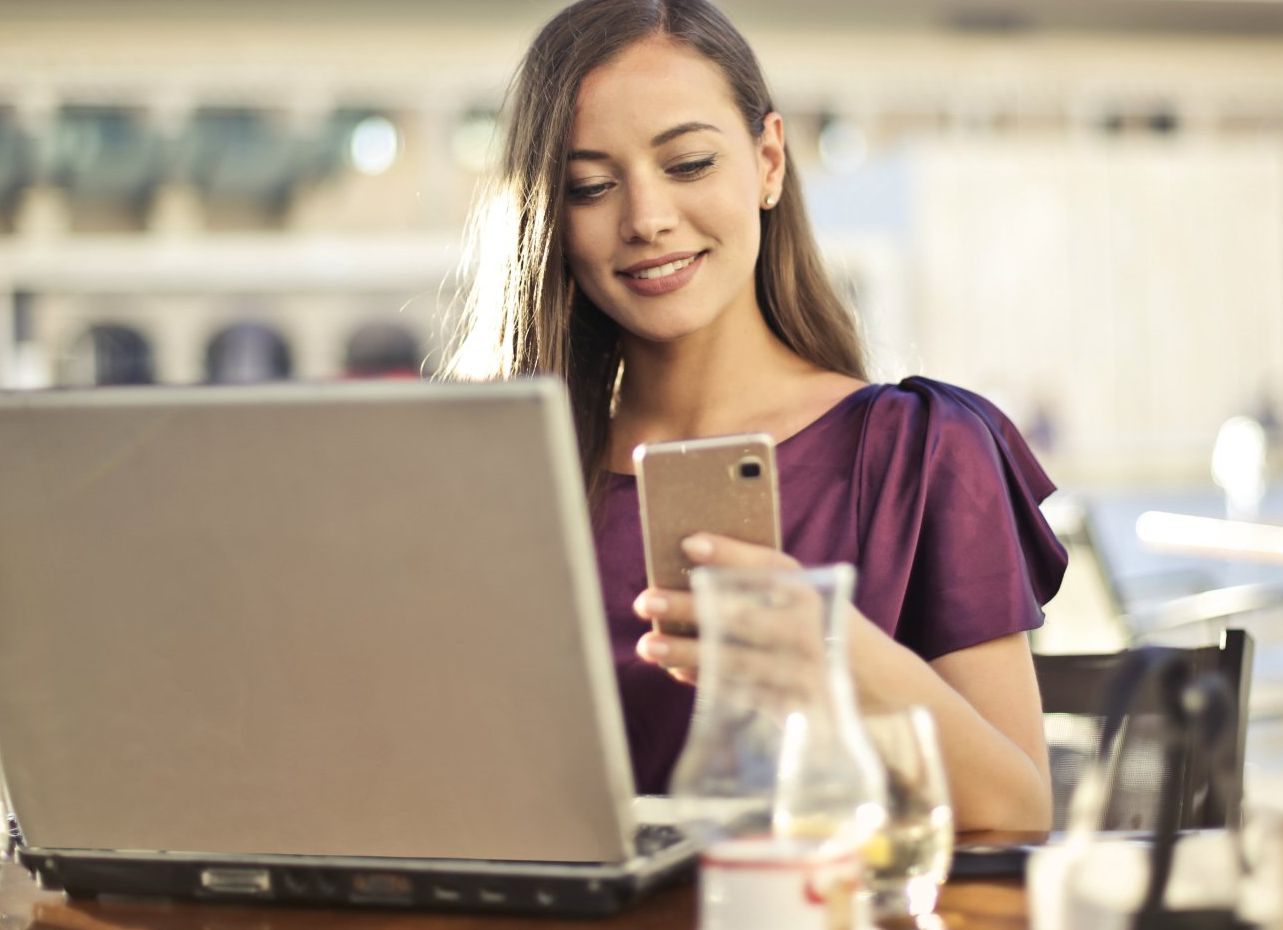 A woman is sitting at a table with a laptop and a cell phone.