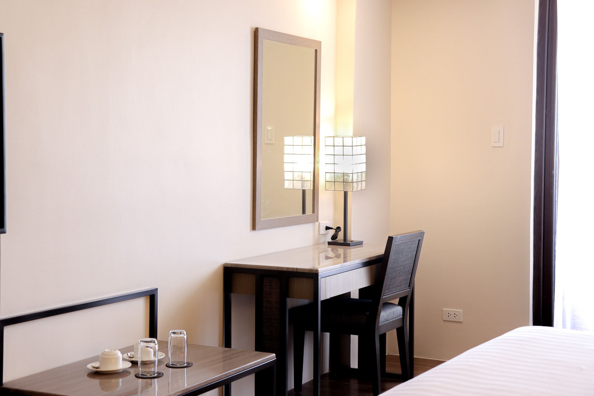 Desk with mirror, lamp, and chair in a hotel room. Table with cups and glasses is on the left. Bright light.