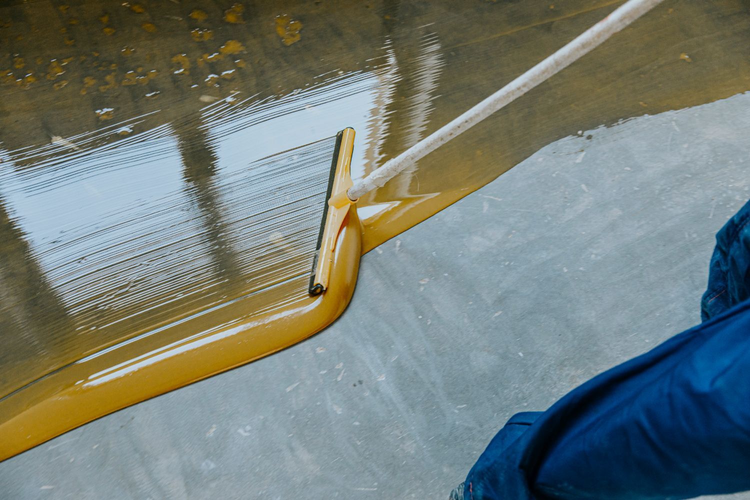 A person is spreading a yellow liquid on a concrete floor with a squeegee.