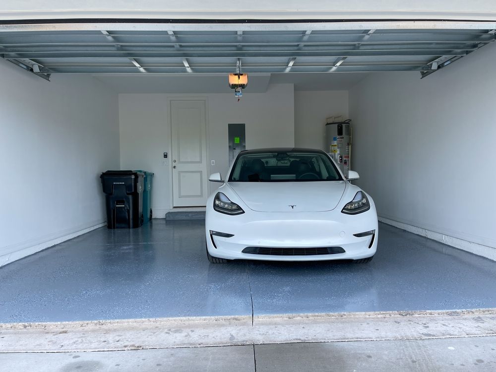 A white tesla model 3 is parked in a garage with the door open.