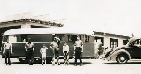 Black and white photo of a group of people standing next to a vintage trailer and car, in front of a building.