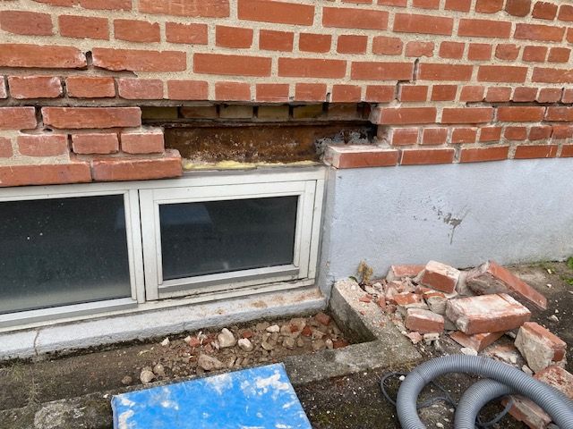 Beskadiget murstensvæg med en manglende sektion over et kældervindue; synlige rester.