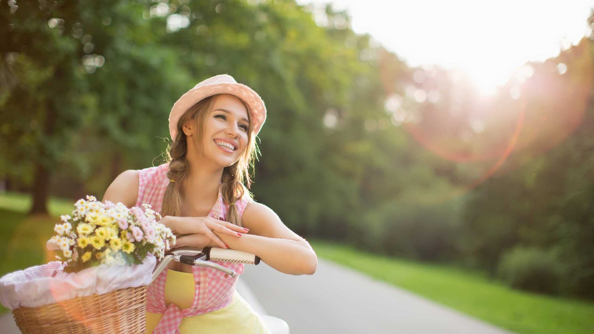 Woman in hat smiles next to bicycle with basket of flowers on a sunny road in a park.