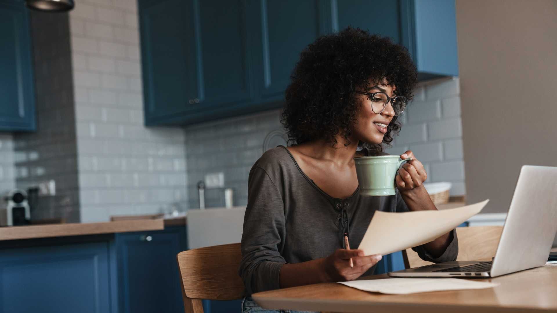 Woman with glasses holding a mug and papers, working on a laptop at a table in a kitchen.