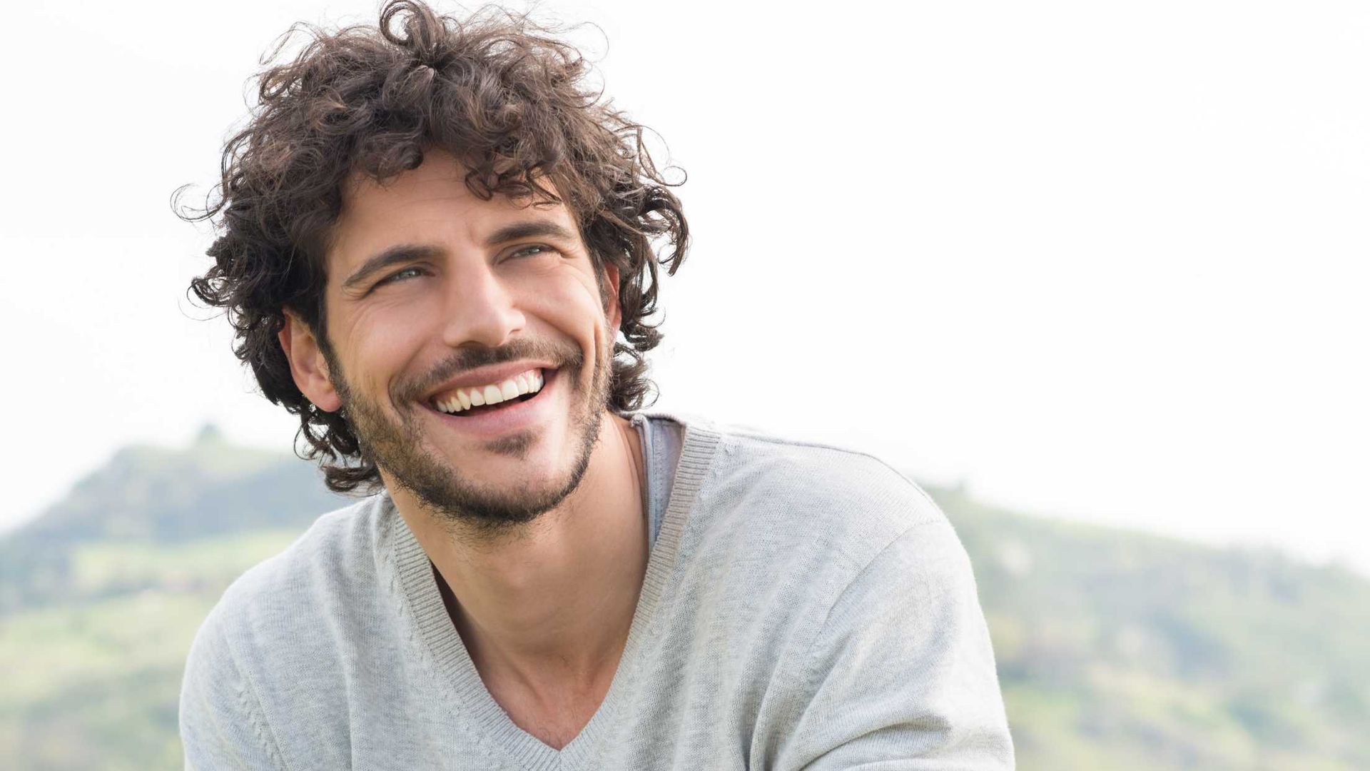 Man with curly hair smiles widely outdoors.