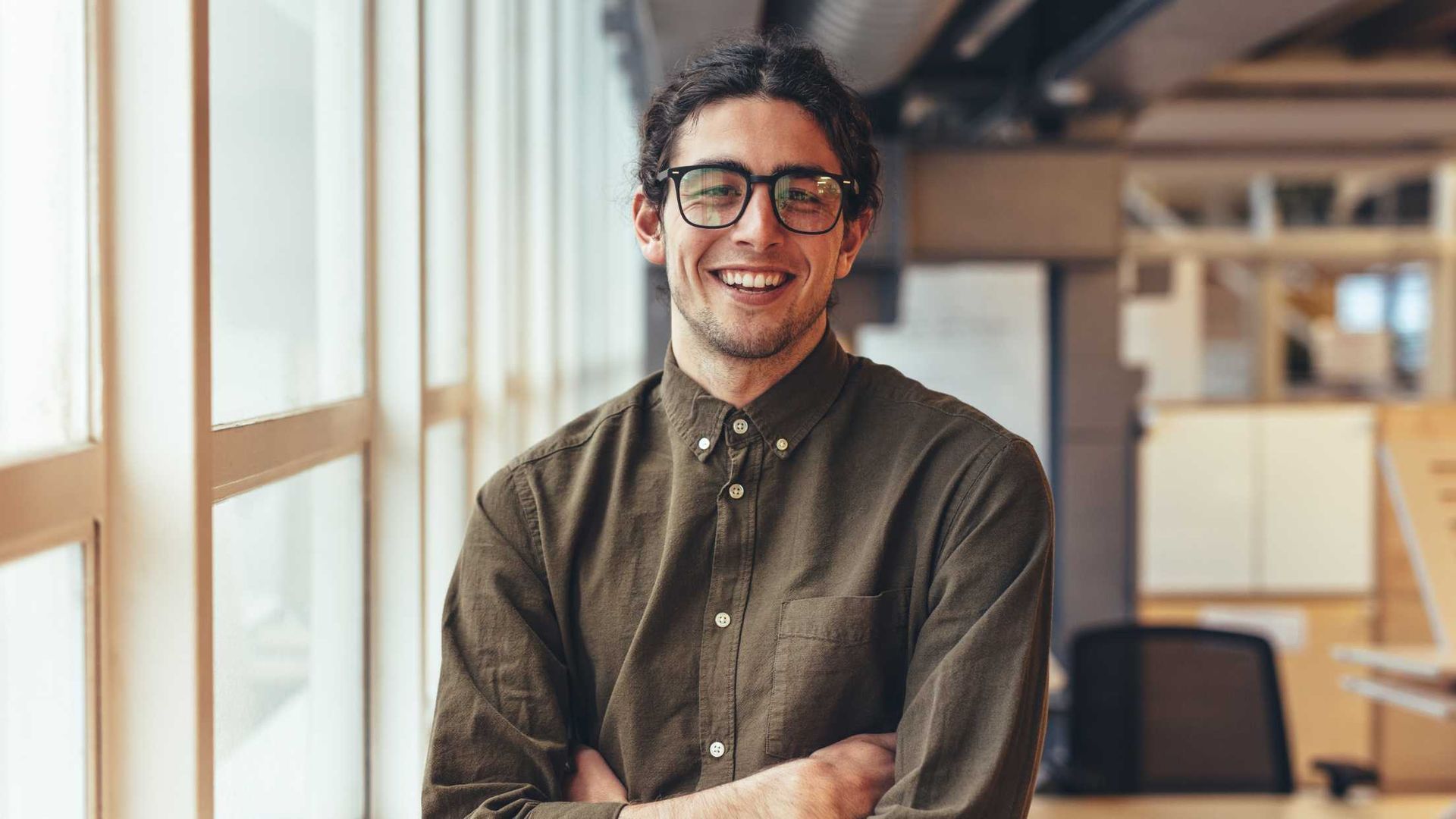 Man with glasses smiling, arms crossed in office setting near a window.