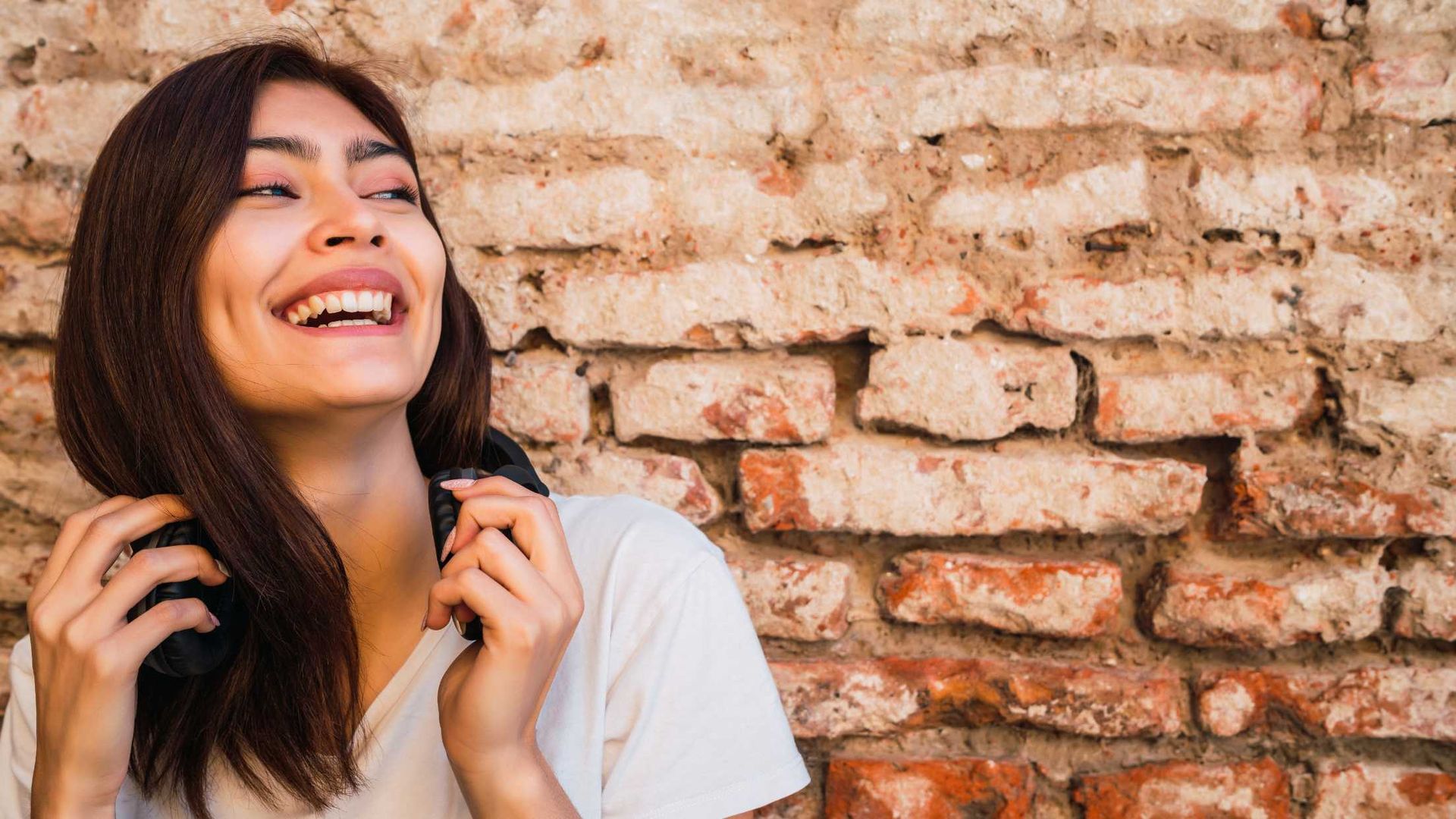 Woman smiling broadly, holding headphones in front of a brick wall.