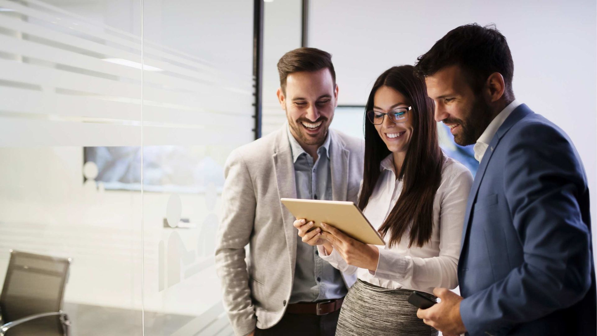 Three business professionals smiling, looking at a tablet in a modern office setting.