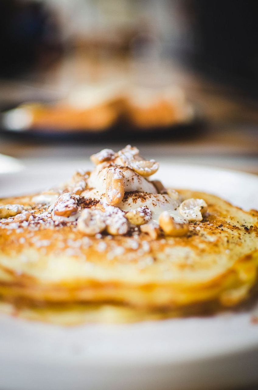 Stack of pancakes topped with cream, nuts, and dusted with powdered sugar, on a white plate.