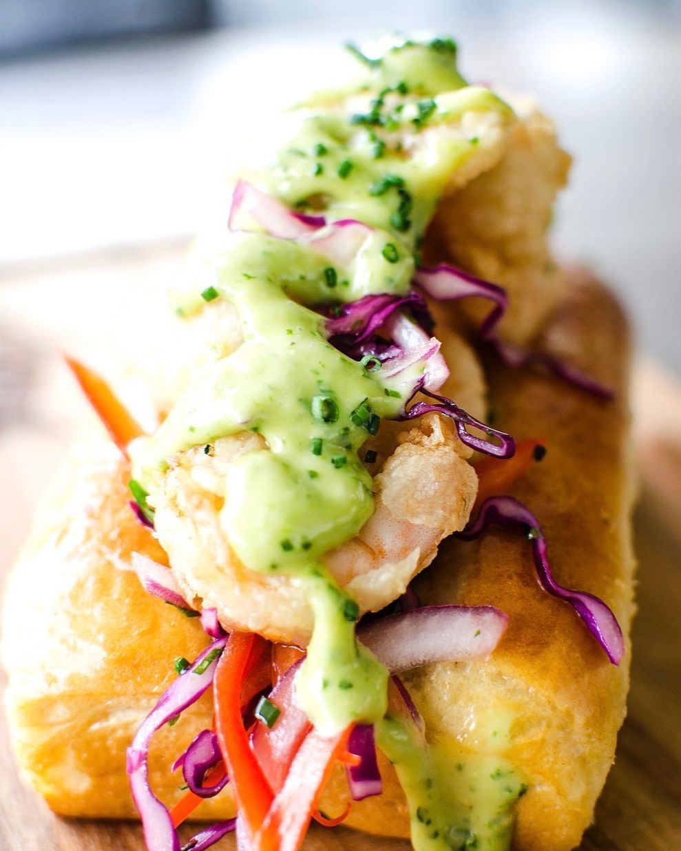 Shrimp roll on a toasted bun, topped with green sauce, red cabbage, and herbs.