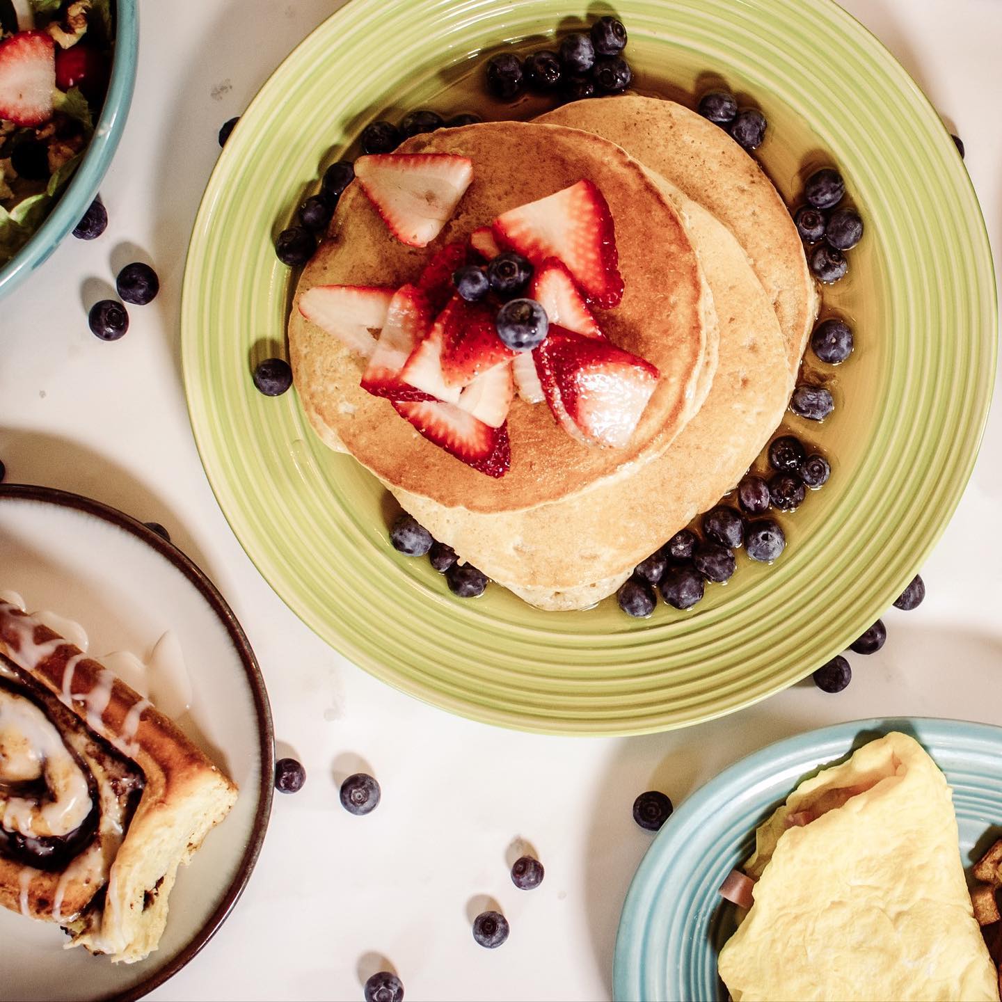 Pancakes topped with strawberries and blueberries on a green plate. Breakfast spread with pastry, salad, and omelet.