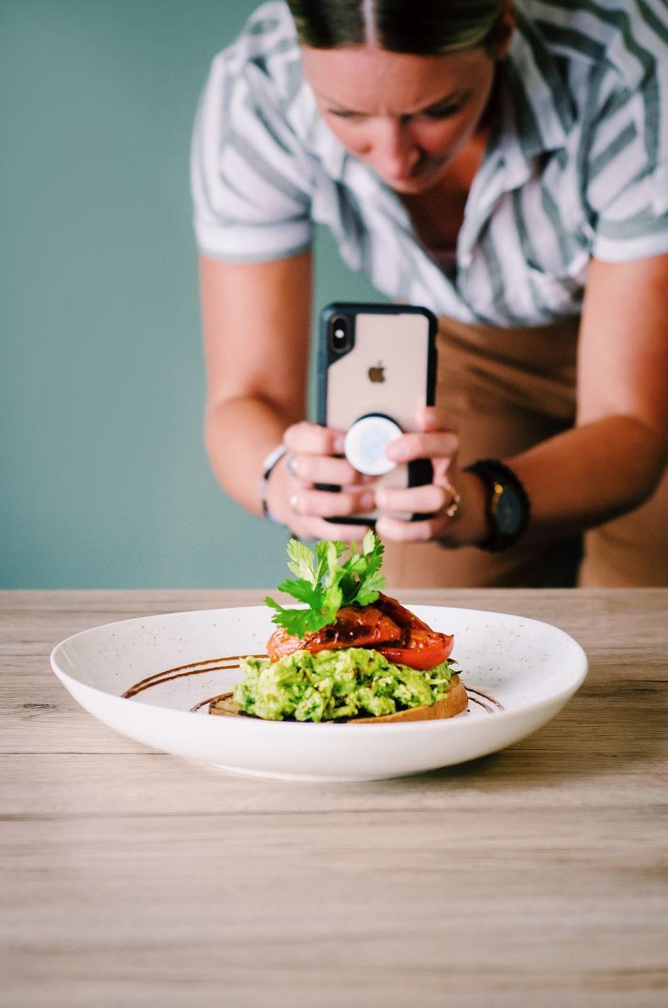 Woman taking a photo of avocado toast on a white plate; light wood table, blue-green wall.