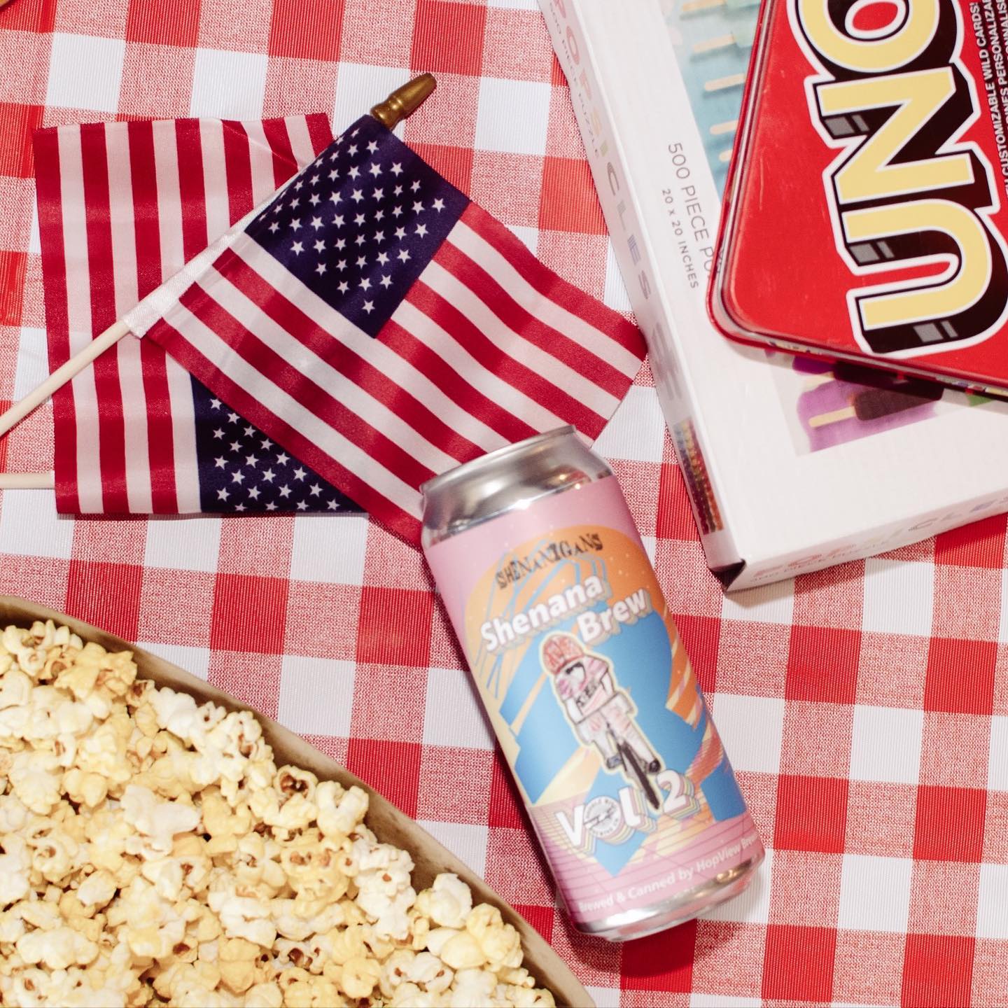 Picnic scene: beer can, American flags, popcorn, and Uno game on a red and white checkered tablecloth.