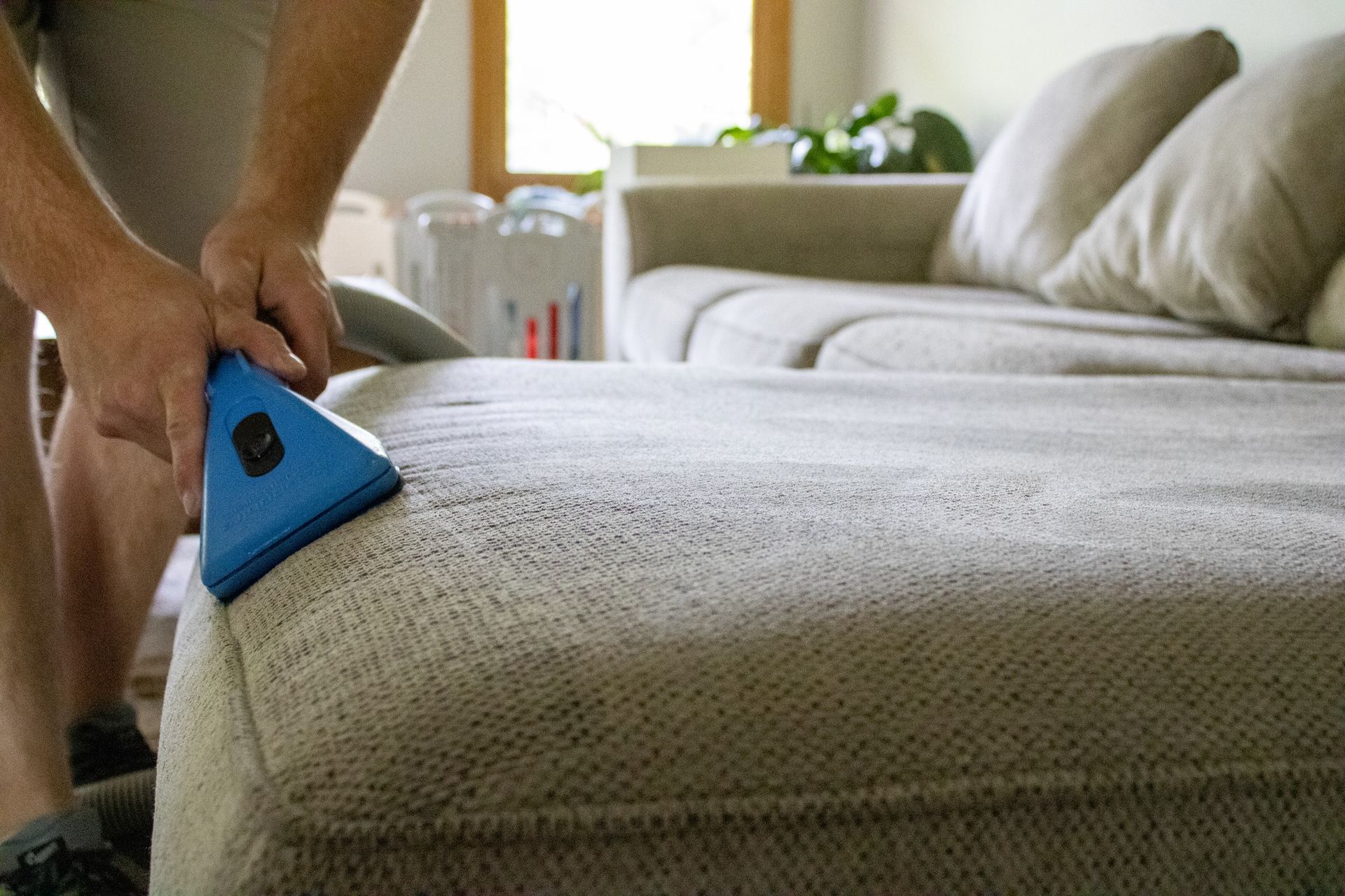 Person cleaning a beige sectional sofa with a blue upholstery cleaner in a living room.