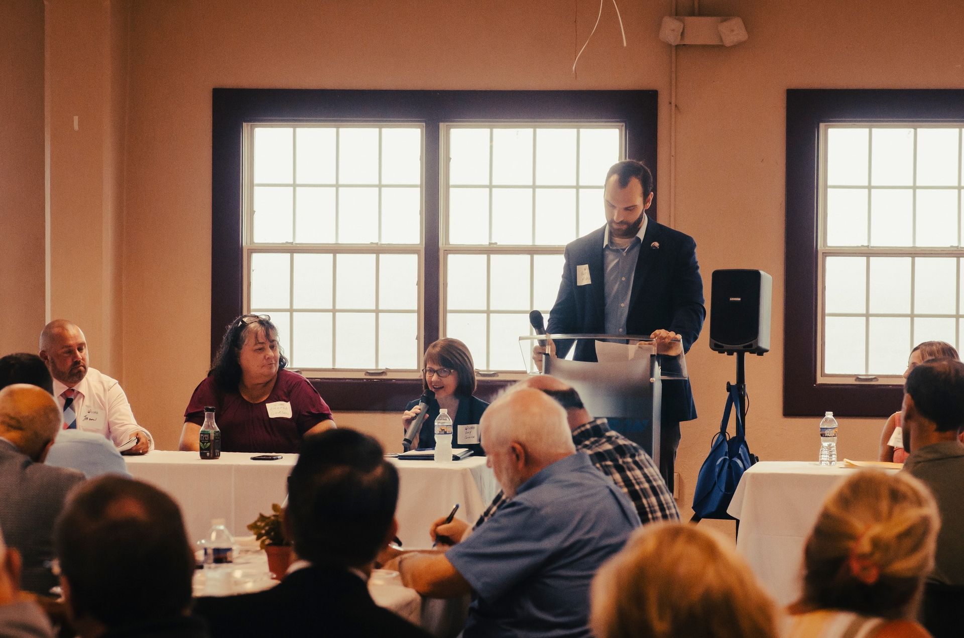 A man speaks at a podium to an audience during a panel discussion; people are seated at a table.