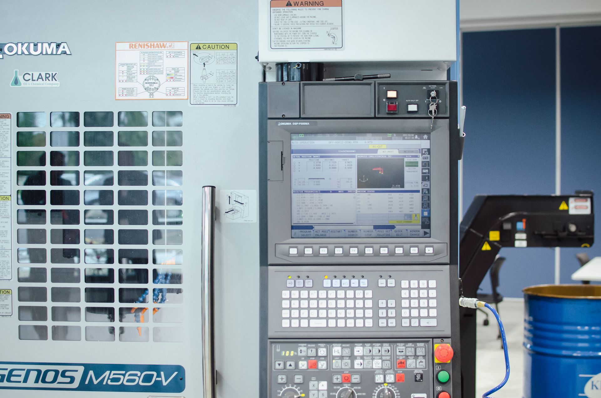 A CNC machine, Okuma Genos M560V, with a control panel and screen displaying machining data.