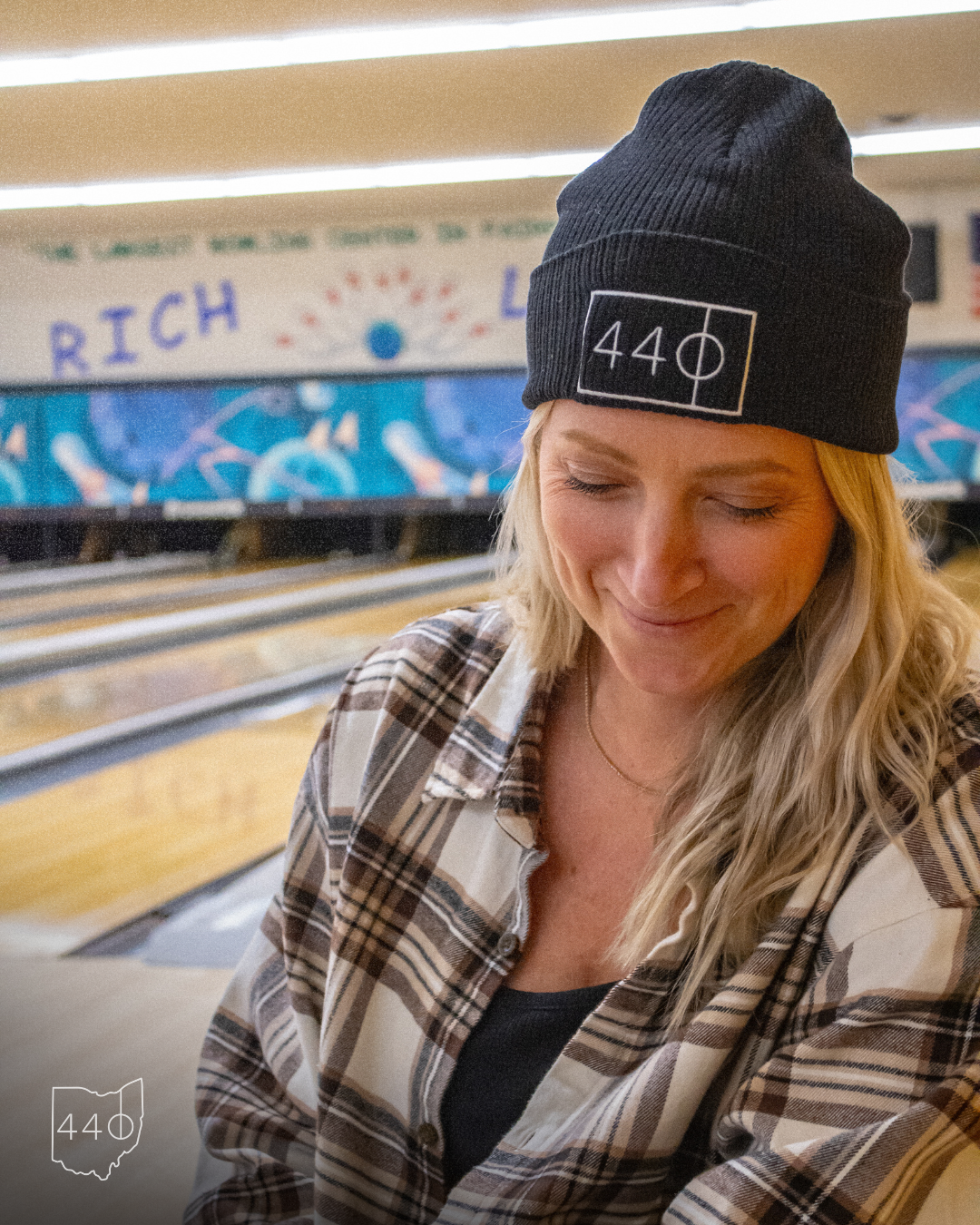 Woman in a black beanie and plaid shirt at a bowling alley.