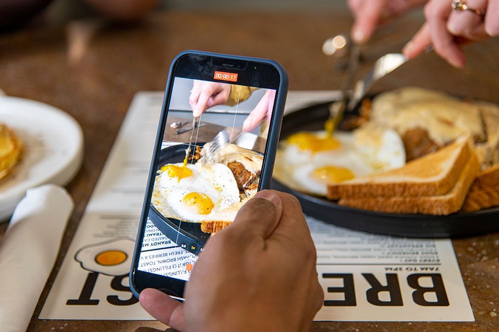 Person films breakfast with a phone. Fried eggs, toast, and hash browns are on a plate on a table.