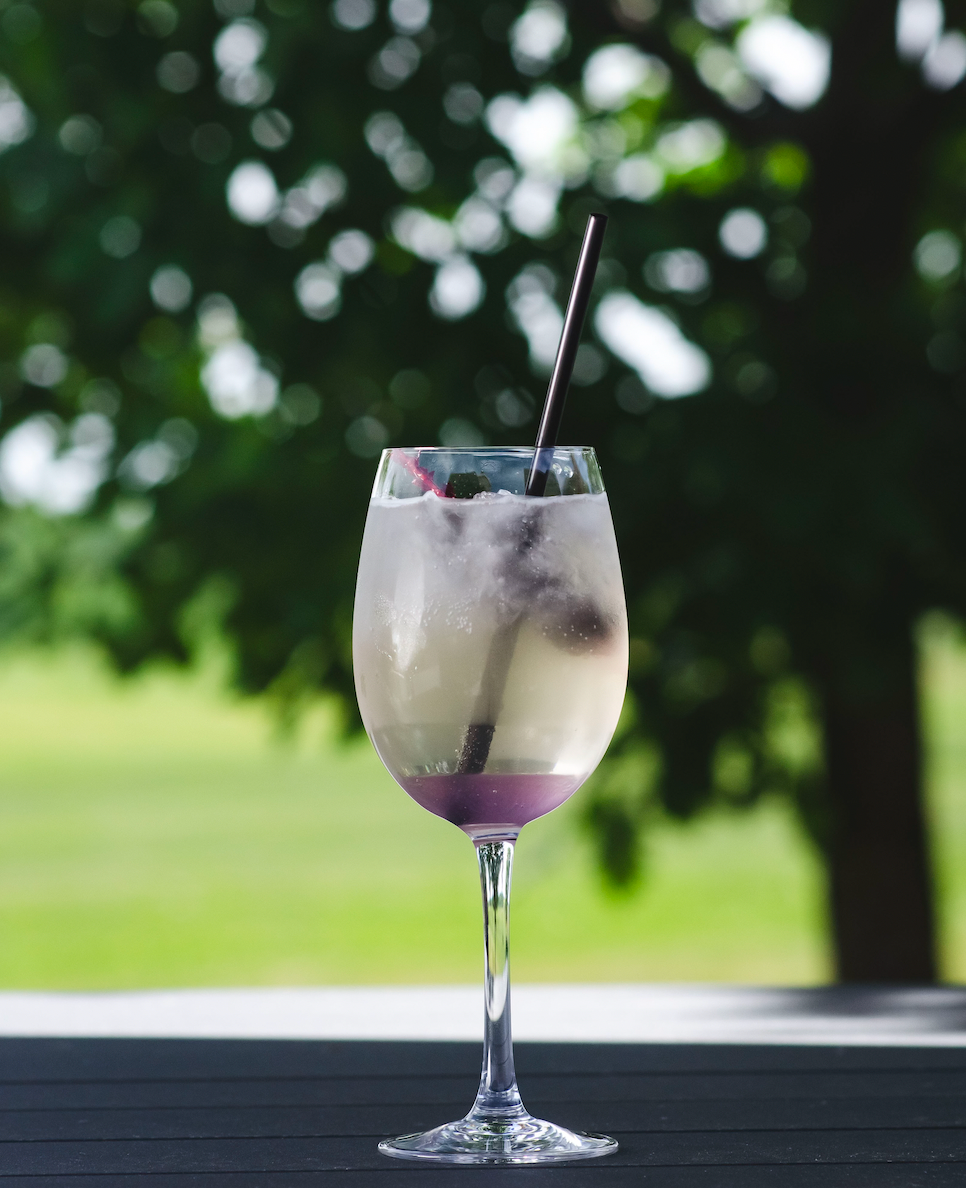 Cocktail in a tall glass with purple layer, straw, on a deck with a green background.