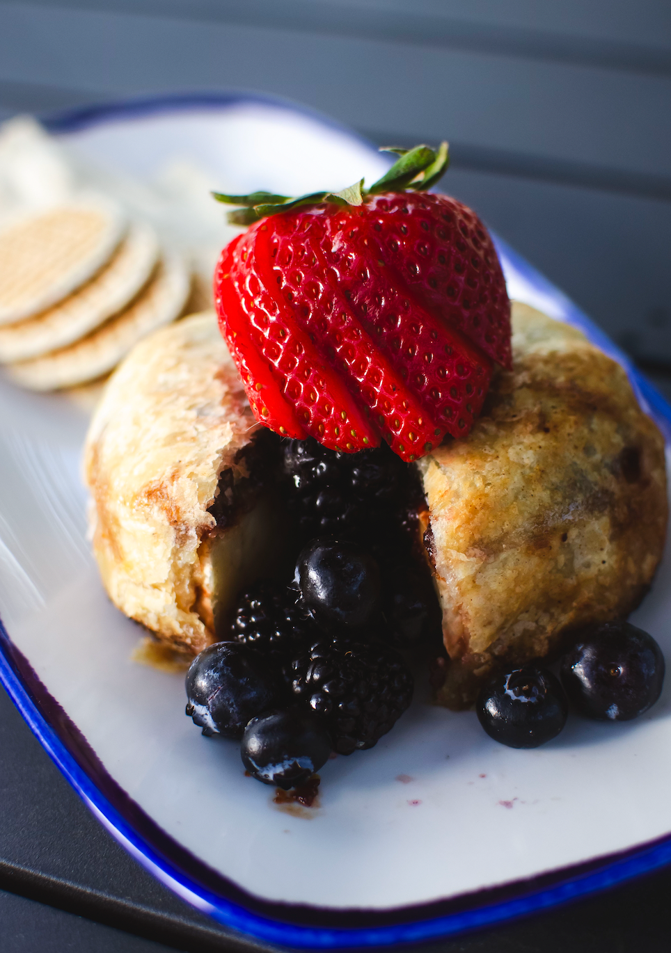 Deep-fried dessert with a berry filling, topped with a strawberry, blueberries, and mini pancakes on a plate.