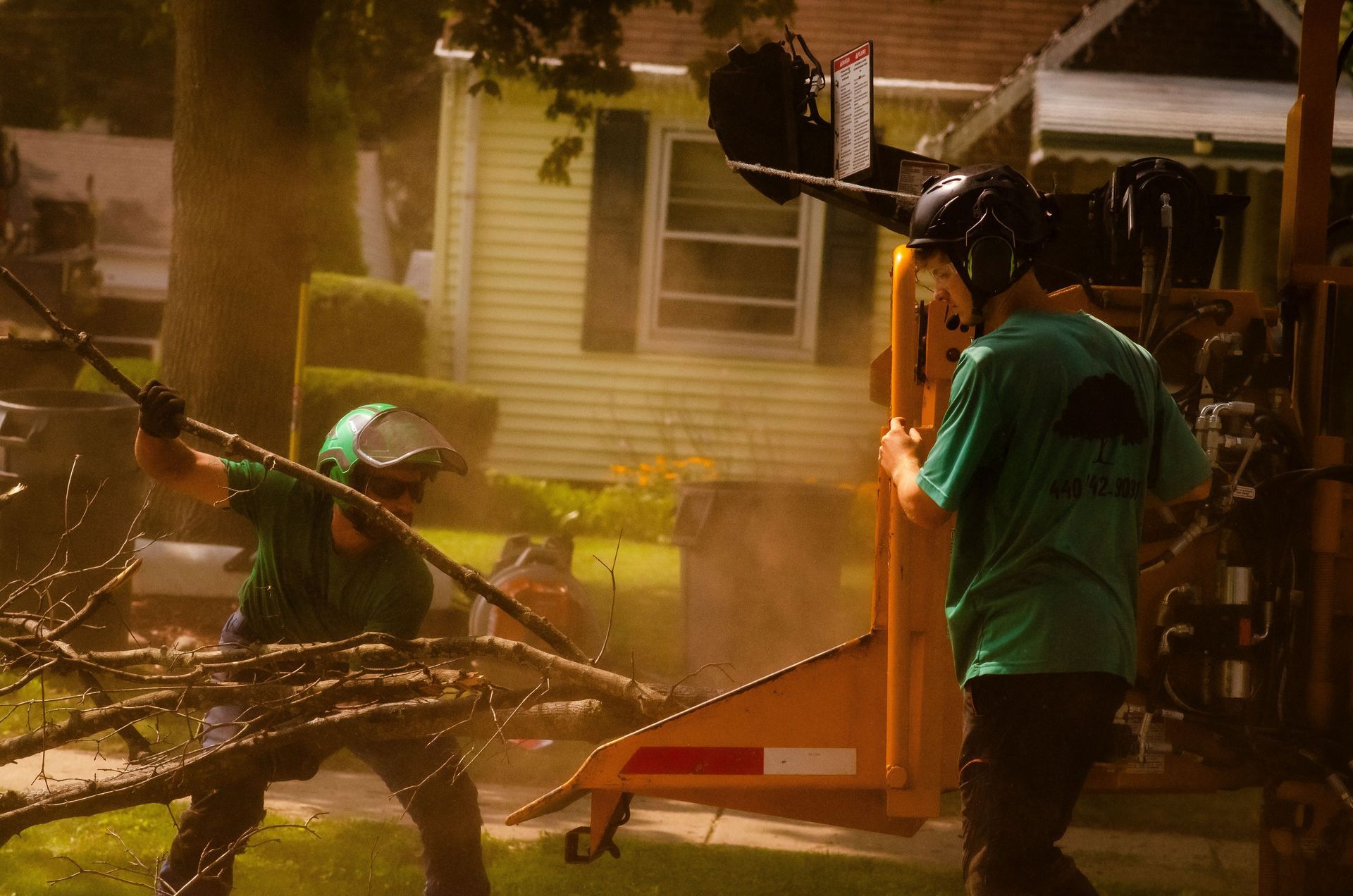 Two tree workers feed branches into a wood chipper in front of a house.