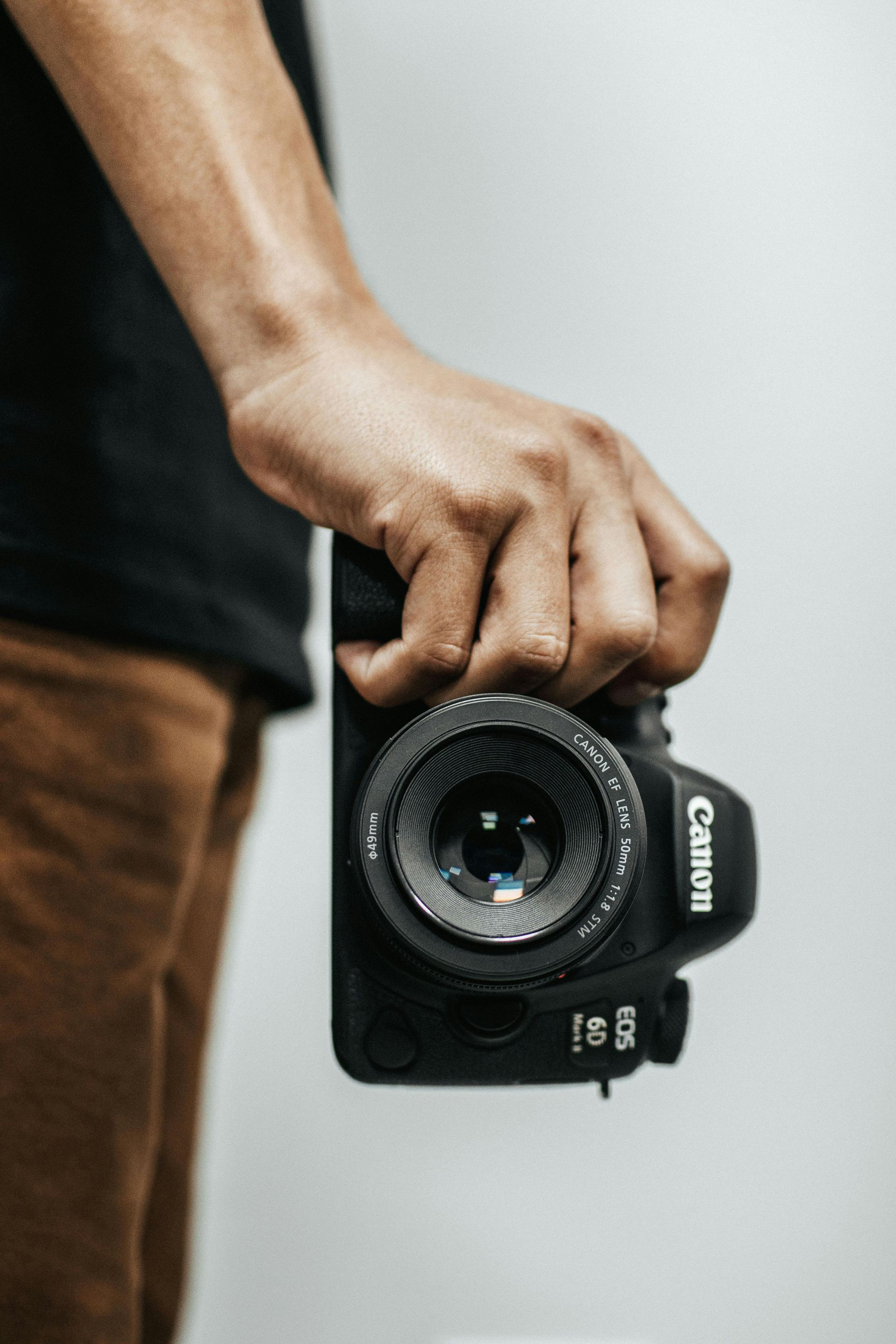 Person holding a Canon DSLR camera. Black camera, tan pants, and black shirt.