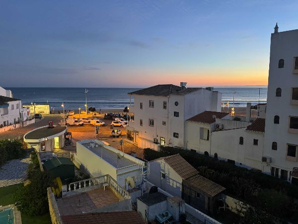 Coastal cityscape at dusk, with buildings overlooking the ocean. Orange and blue sky, street lights on.