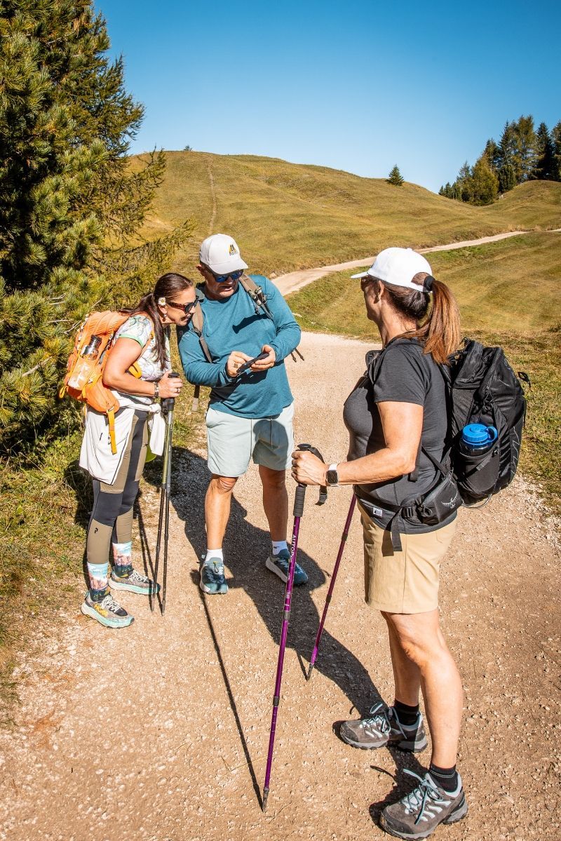 Three hikers on a trail consult a map on a phone. Sunny day, green hills in background.