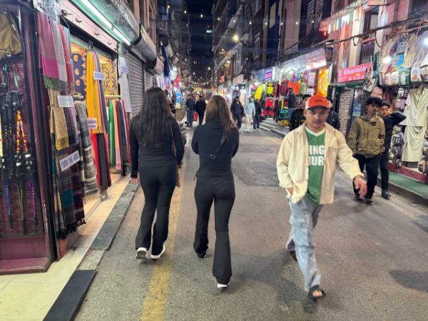 People walking on a narrow street lined with shops at night. Fluorescent lights illuminate the stores.