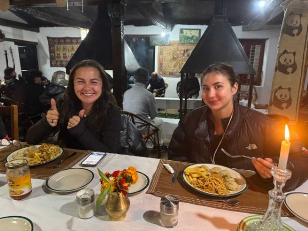 Two people seated, smiling at a table, eating in a warmly lit restaurant. One gives a thumbs up.