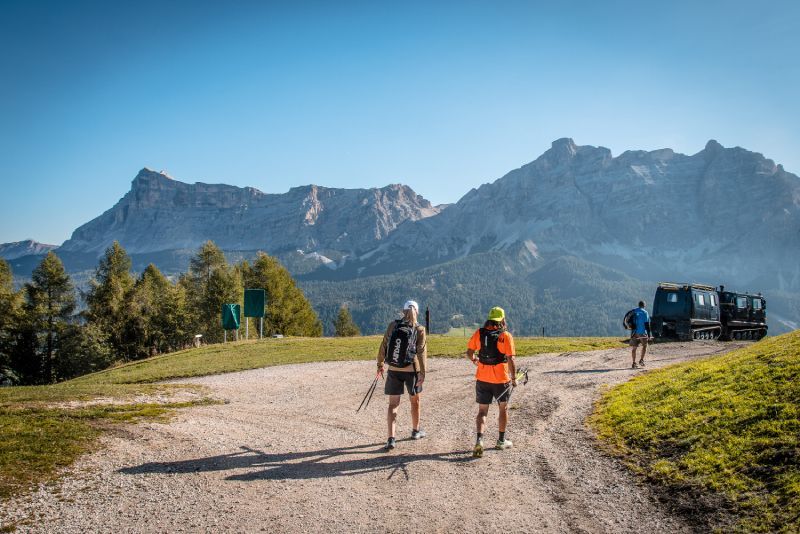 Two runners hiking towards mountains on a dirt path, with a support vehicle and blue sky.