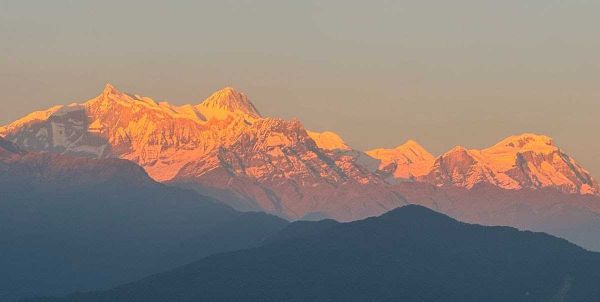 Snow-capped mountain range illuminated by the golden light of sunrise or sunset, with rolling hills in the foreground.