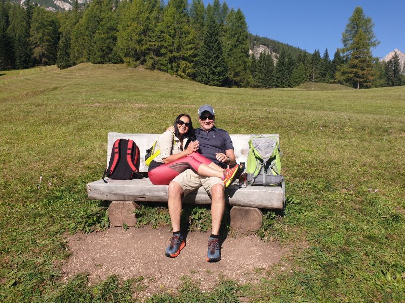 Couple seated on a bench in a grassy field, mountains in the background.